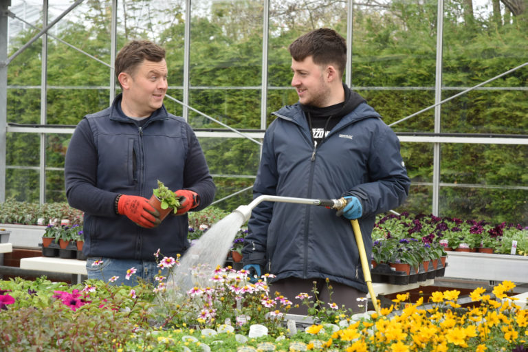 staff and student in the green house looking at each other. the student is watering the plants