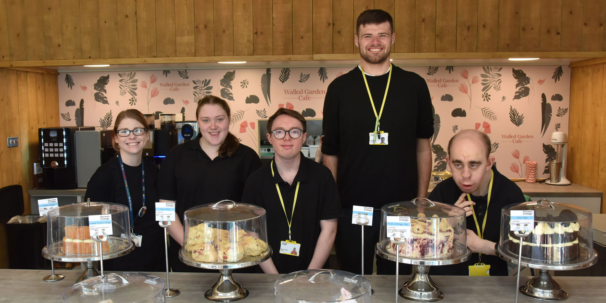 students and staff standing behind a counter in the cafe. the counter has lots of cakes on it.