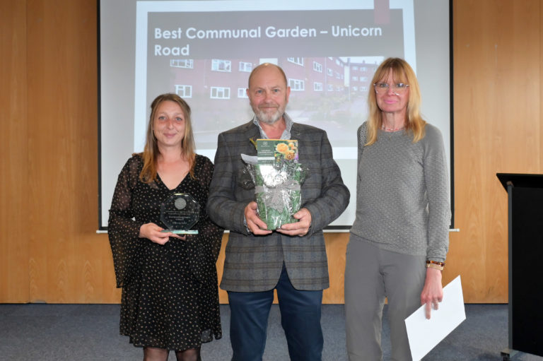 Commercial manager Pete holding a potted rose stood with two women holding a certificate and trophy in front of a screen which says 'Best Communal Garden - Unicorn Road.