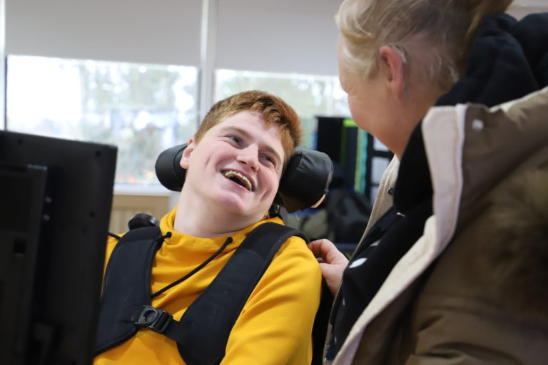 PMLD student in wheelchair laughs as he's supported to use communicative technology by a member of staff