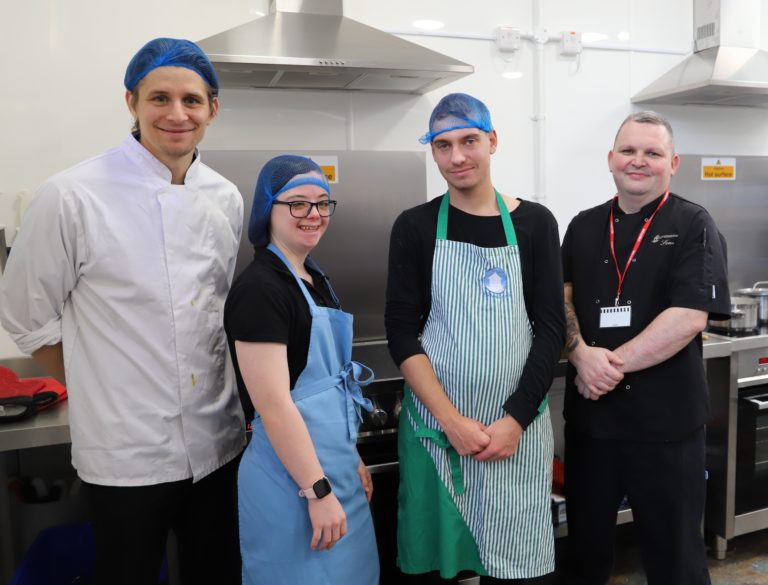 Teacher George, students Helen and Kyle and chef Sean, wearing aprons and hairnets, stand in front of the cooker.