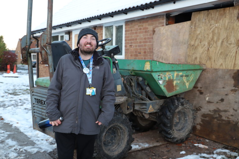 Student Brendan stands in front of one of the bungalows being refurbished which is boarded up and has machinery outside it.