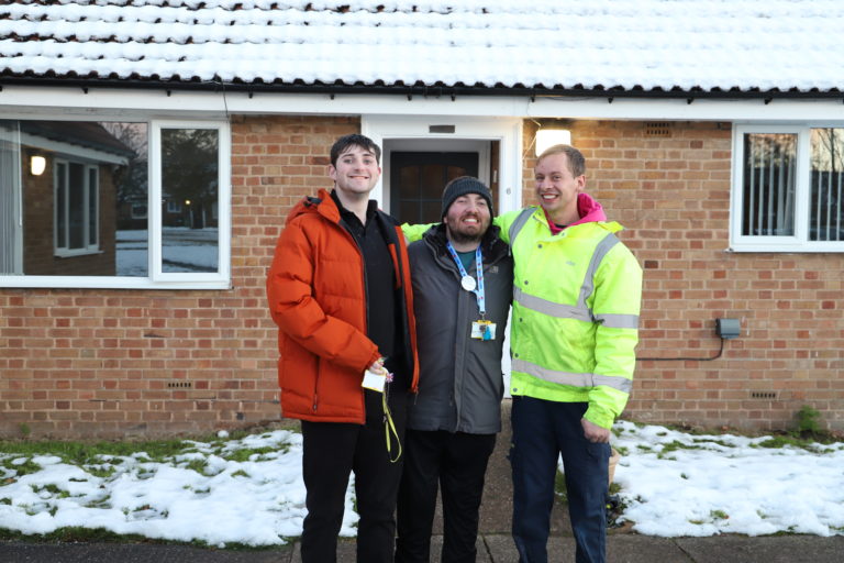 Bryon, Brendan and James stand outside their bungalow with arms around each others shoulders, wearing coats, in the snow.