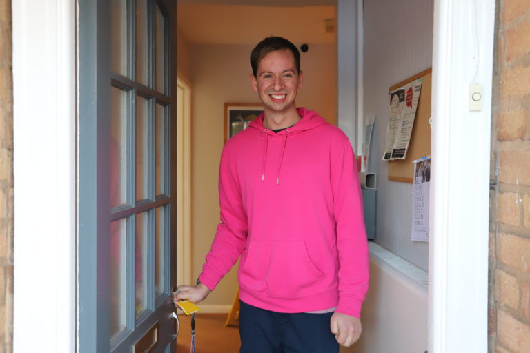 Student James, wearing a pink hoodie, opens the door to his bungalow with a smile.