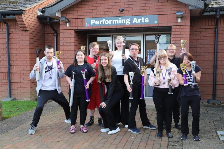 Opal dressed in black, with eight Performing Arts students holding trophies they've won, in front of the Performing Arts building.