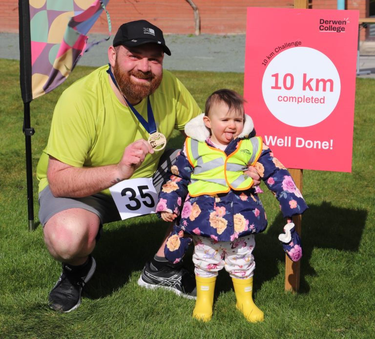 Runner David with toddler daughter in high-vis jackets stood by the 10km finish sign