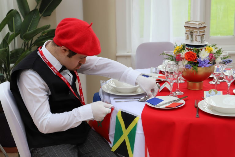 A student dressed in a red beret with white gloves sets a table adorned with flags of Europe with red and blue table cloth and Arc de Triomphe centrepiece.