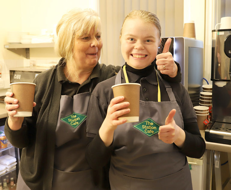 Supervisor Teresa and student Arielle, wearing grey aprons with The Station Cafe green logo, smiling and holding paper cups, with thumbs up.