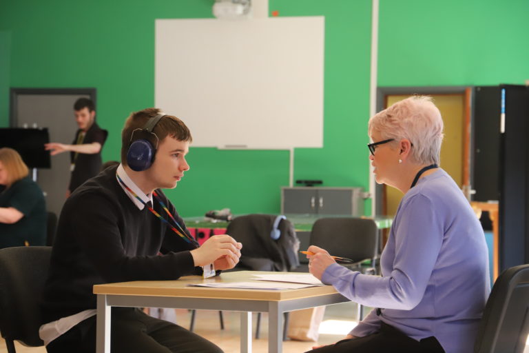 A student wearing ear defenders is sat at a table opposite a woman who is interviewing him.