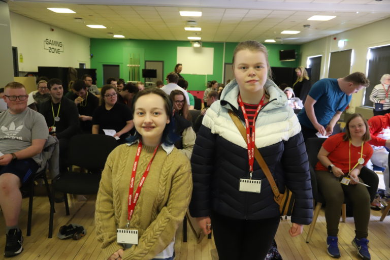 Two female graduates wearing visitors badges stand in front of a packed audience of seated students.